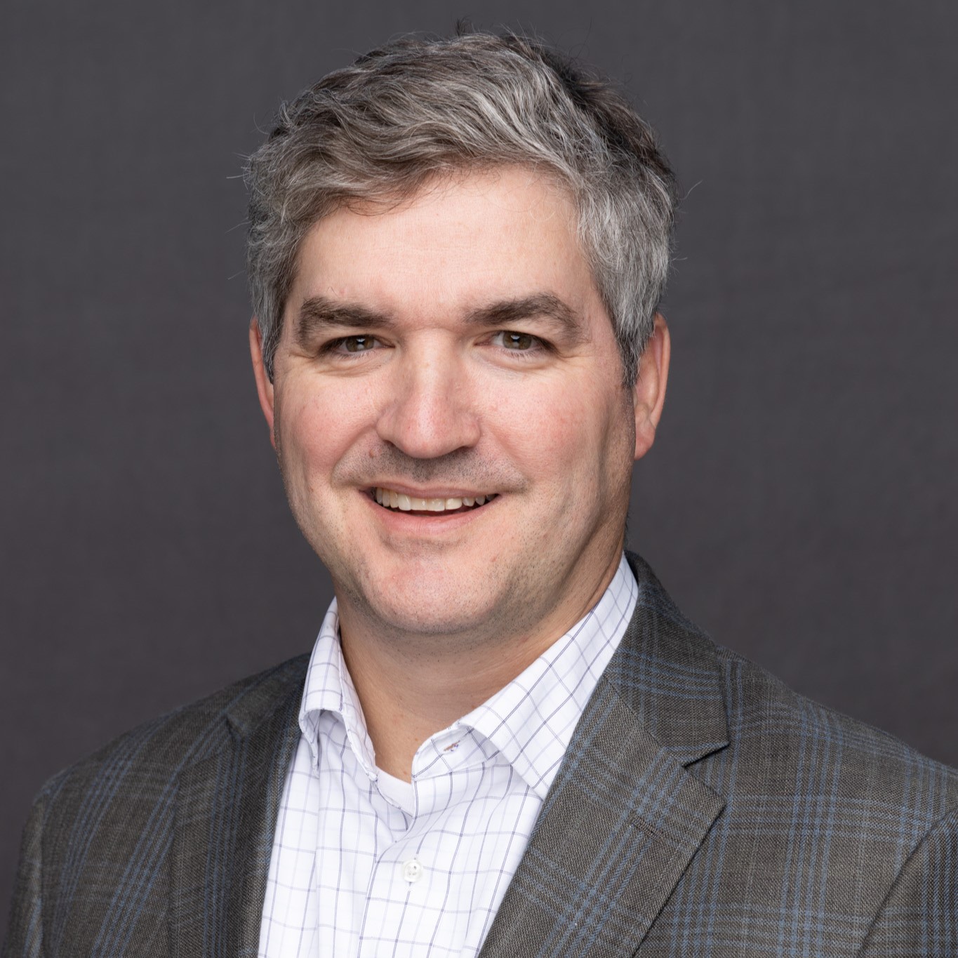 Will Landry headshot. He is wearing a plaid suit jacket and is in front of a dark grey background. 