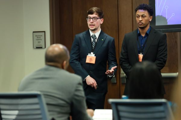 Two students in dark suits present in front of judges. 